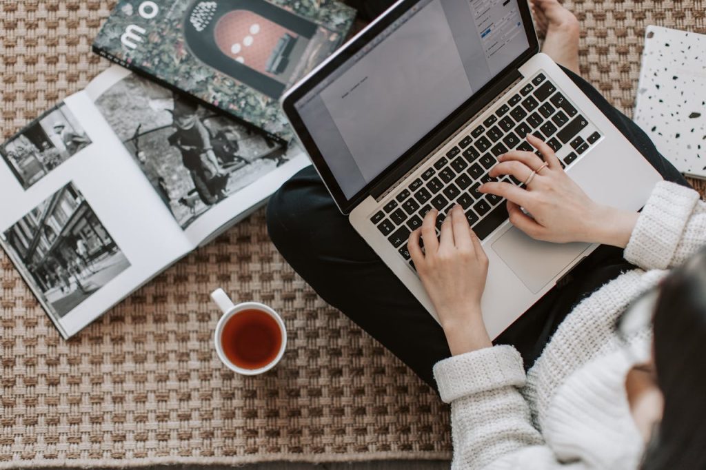 Overhead view of a woman typing on a laptop at home, surrounded by magazines and a cup of tea on the floor.